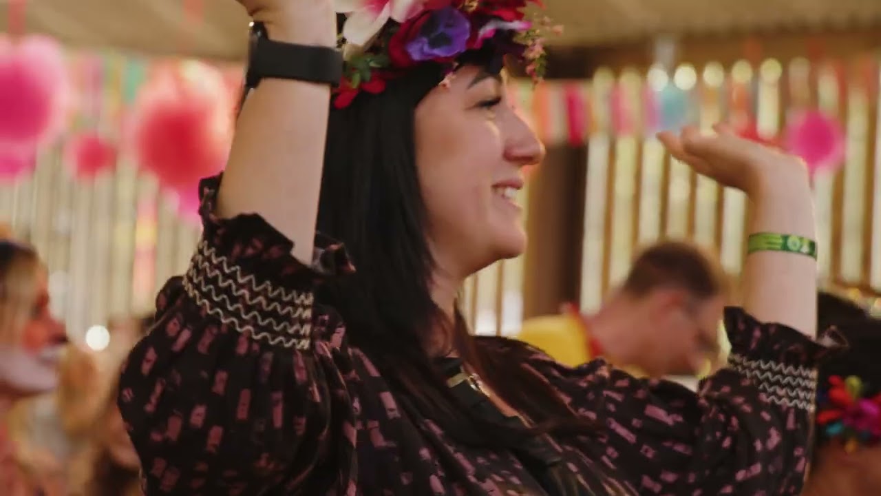 A headshot of a woman in a floral head dress. She has her arms raised and is looking away from the camera