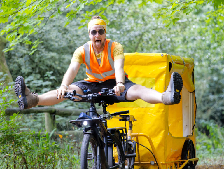 Man riding a bike through the woods with his legs out to the side, towing a yellow trailer