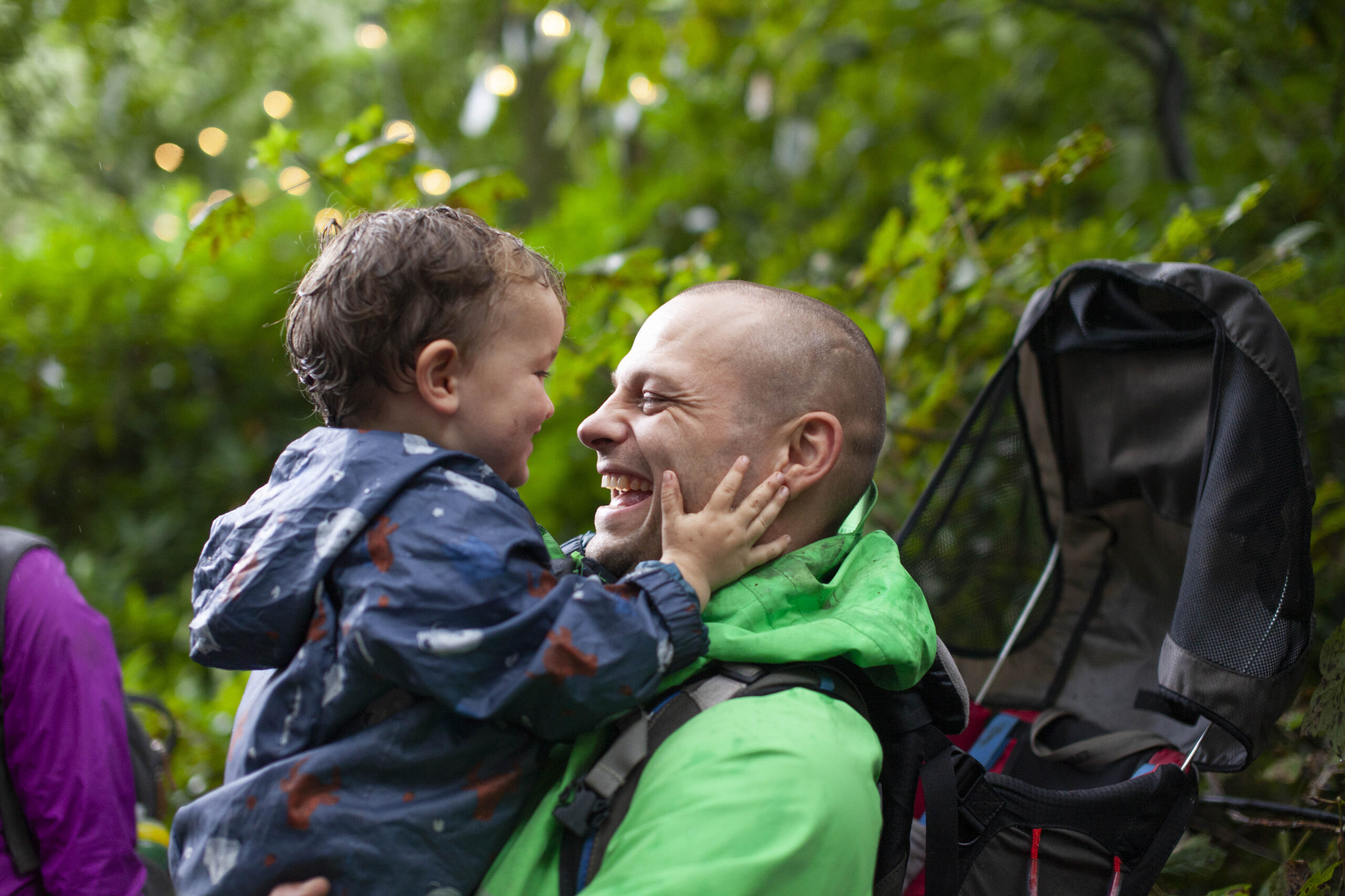 A dad holds his smiling little boy. They are both wearing waterproof coats and are outside in a woodland